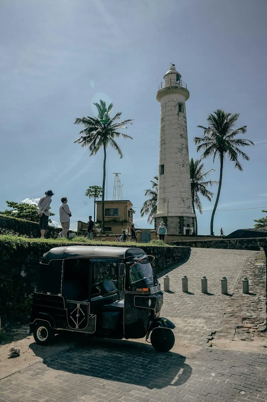 Two blue tuk-tuks driving along a sunny Sri Lankan coastal road lined with palm trees and guesthouses - the quintessential self-drive experience in Sri Lanka