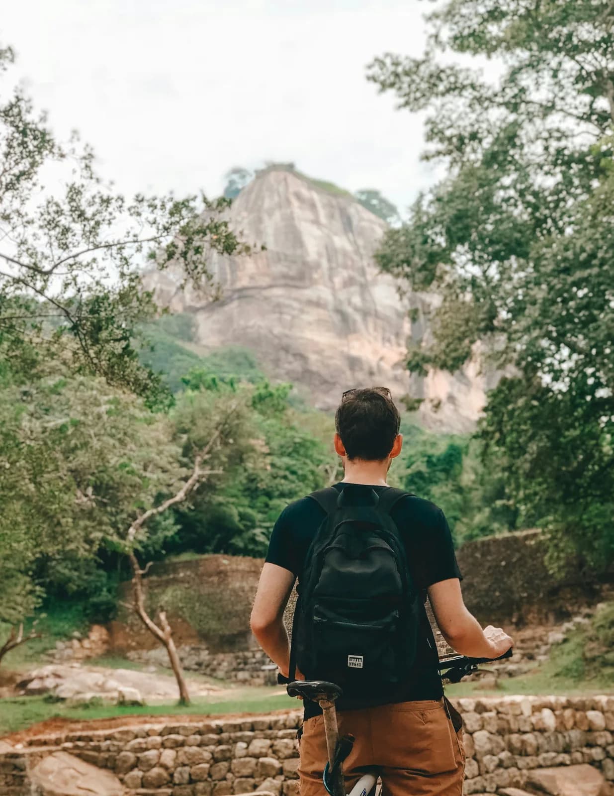 A traveller with a backpack cycling through the ancient ruins of Polonnaruwa, Sri Lanka, with a large rock formation visible through green forest