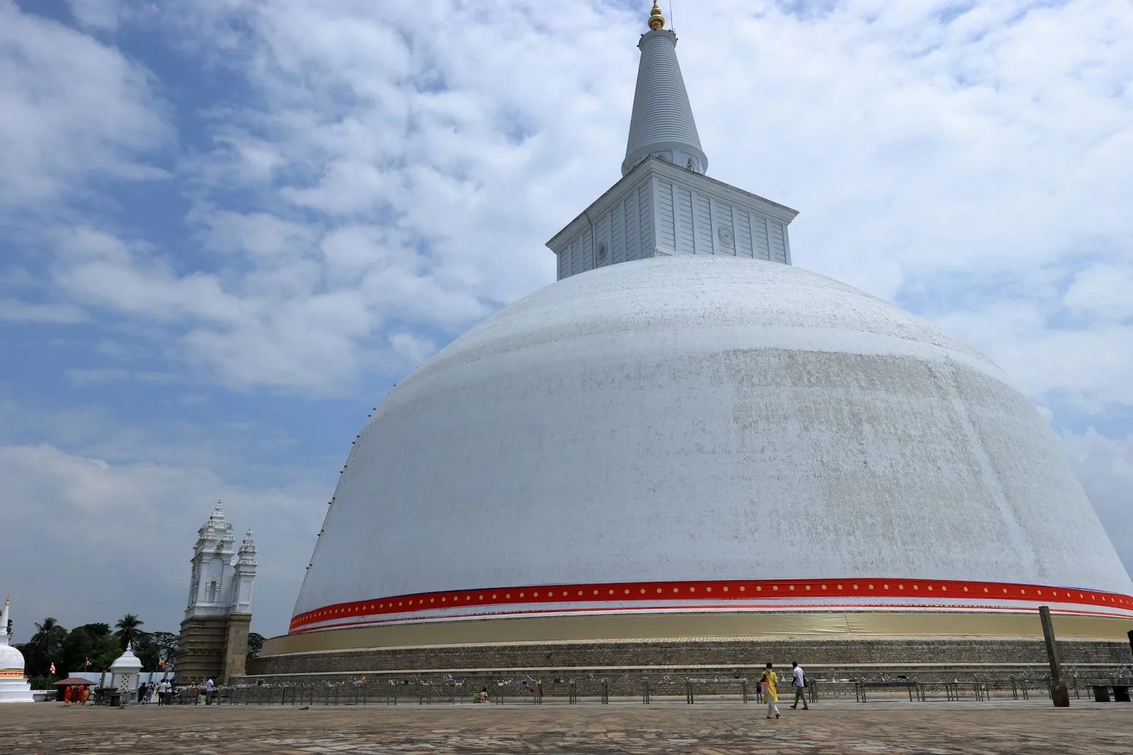 The white Ruwanwelisaya stupa at Anuradhapura rising above the treetops with a clear blue sky and Buddhist devotees walking the perimeter path