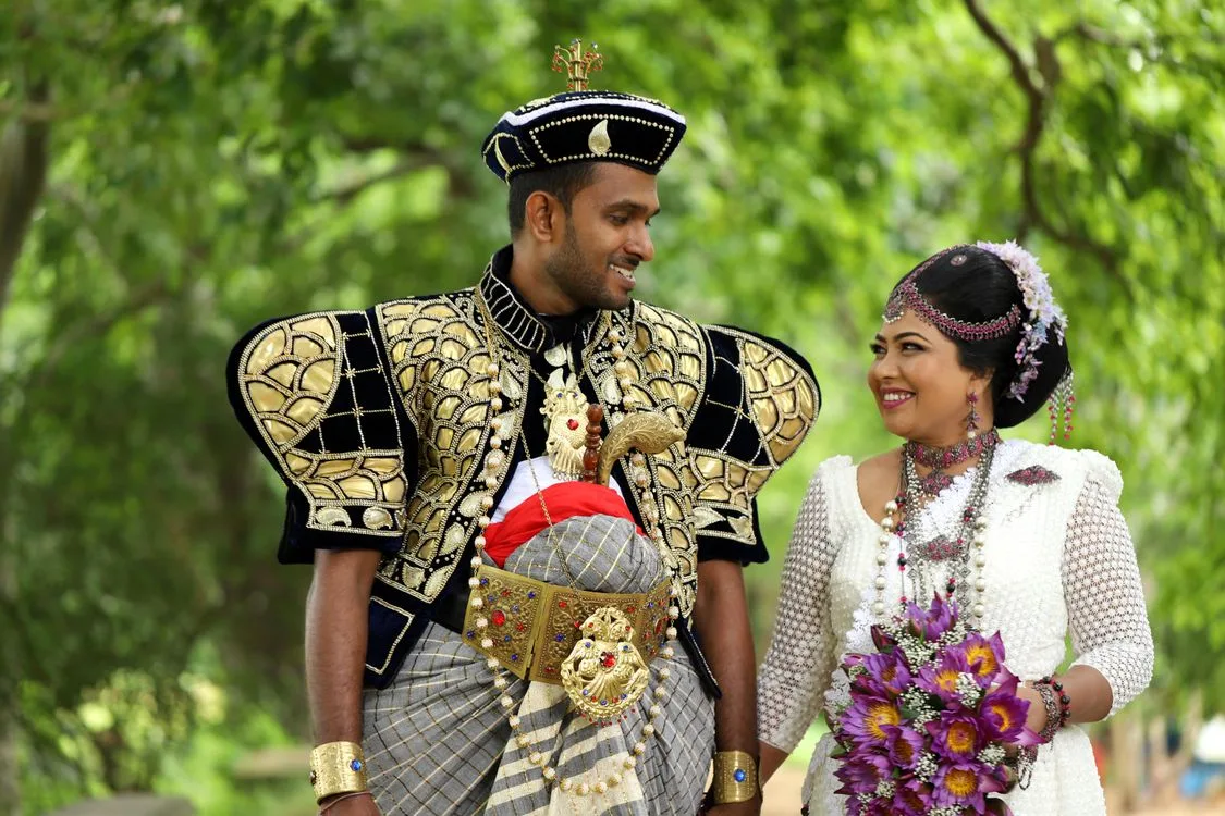 A Sri Lankan couple in ornate traditional wedding attire set against a lush tropical green backdrop - the island's natural scenery creates a distinctive visual identity for destination weddings