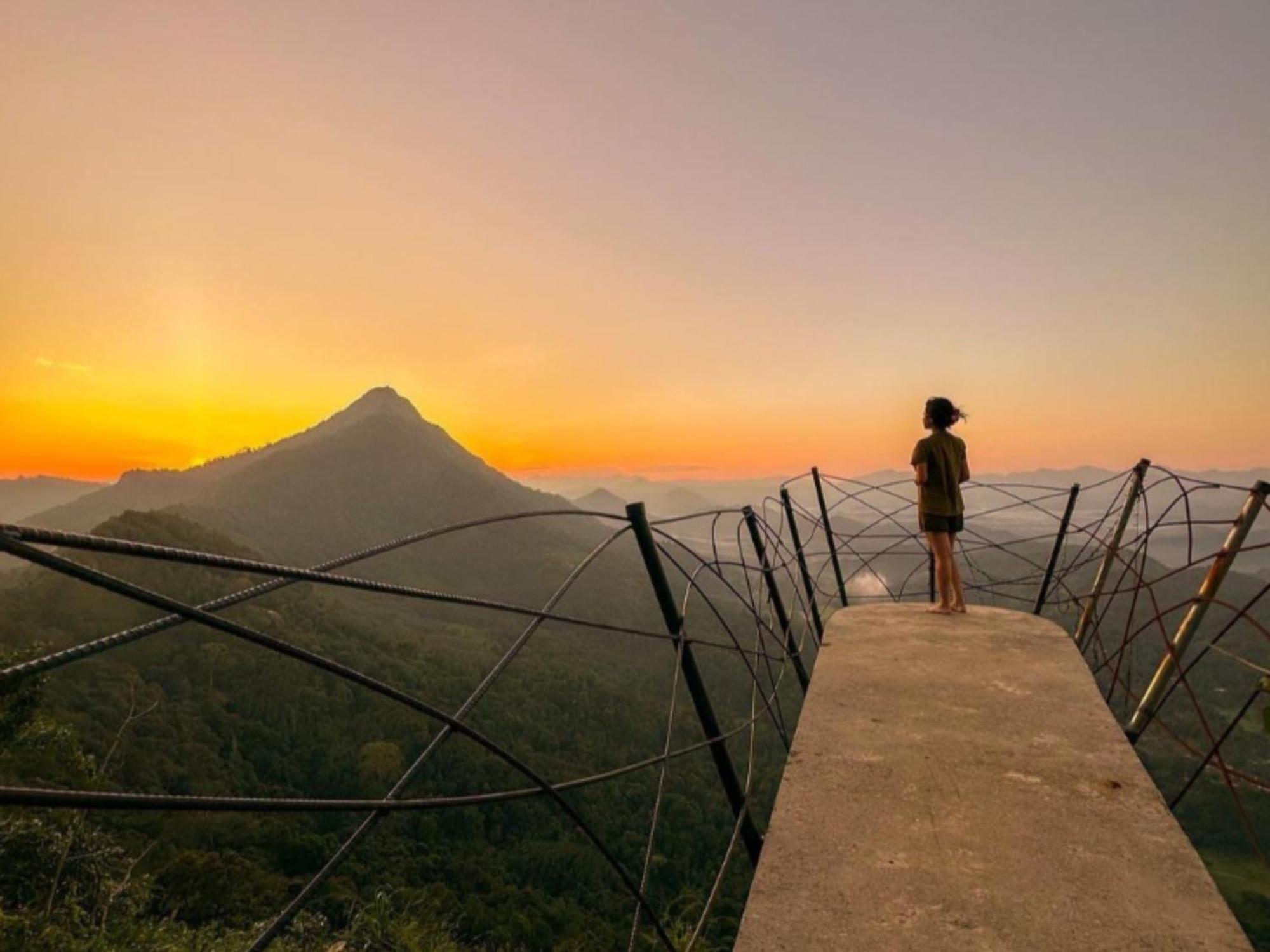 A person standing alone on the Greenacres viewpoint platform at sunset, silhouetted against a vivid orange and pink sky with a peaked mountain and misty valleys below