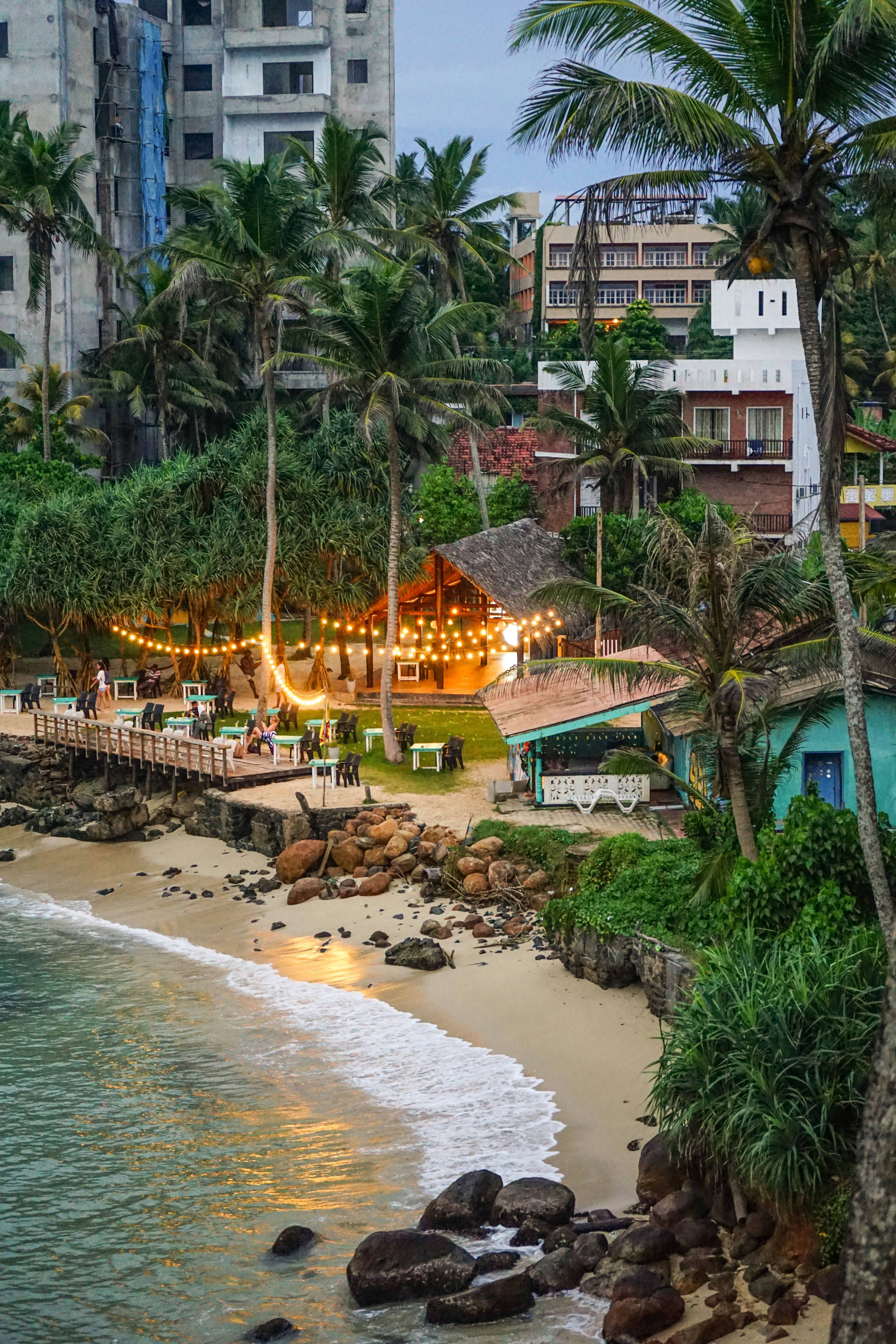 A small beach cove in Colombo at dusk with fairy-light restaurant, palm trees, and gentle surf