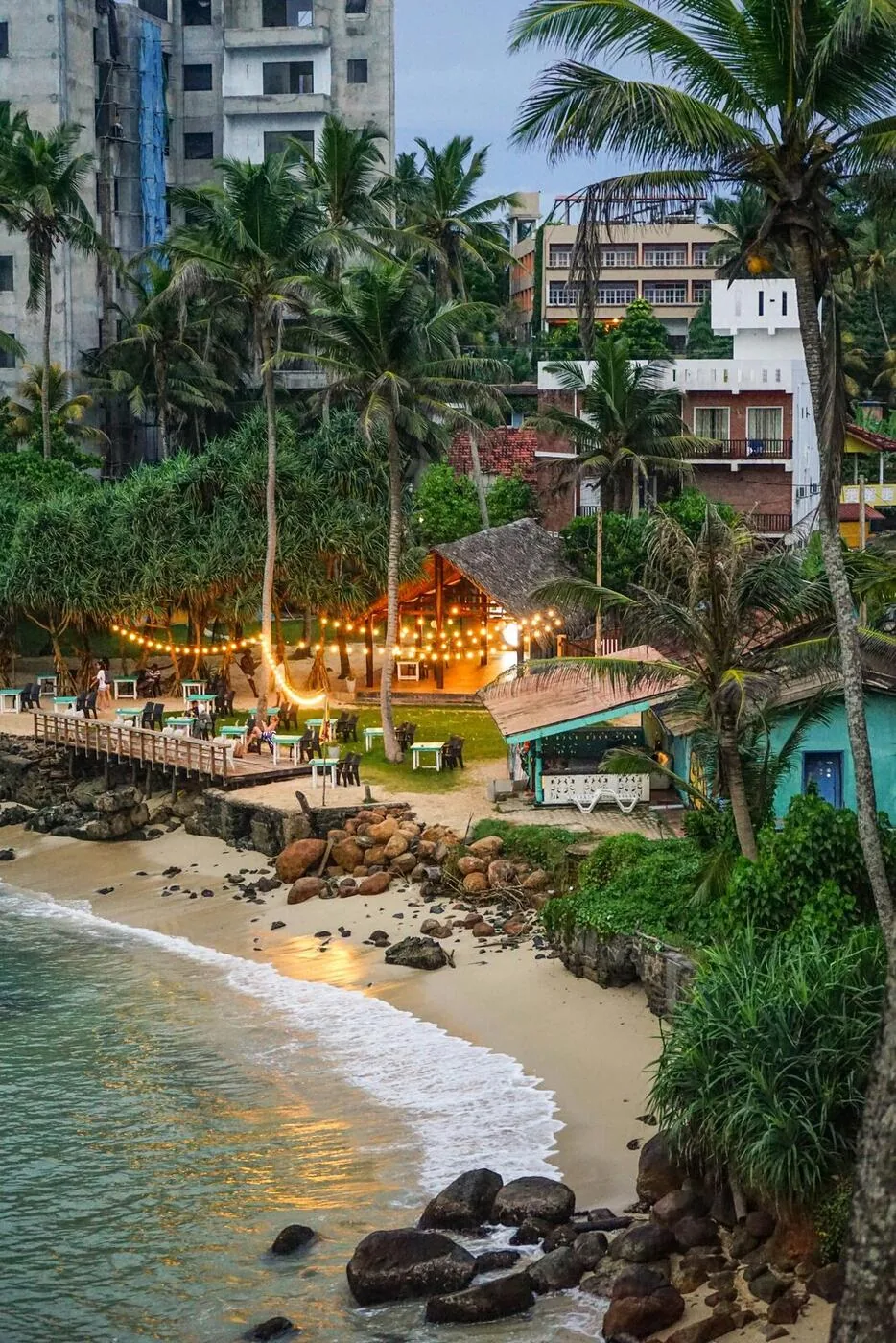 A small beach cove in Colombo at dusk with fairy-light restaurant, palm trees, and gentle surf