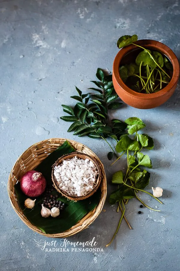 Fresh ingredients for Kola Kanda laid out on a wooden surface including gotu kola leaves, curry leaves, red rice, fresh coconut, garlic and shallots