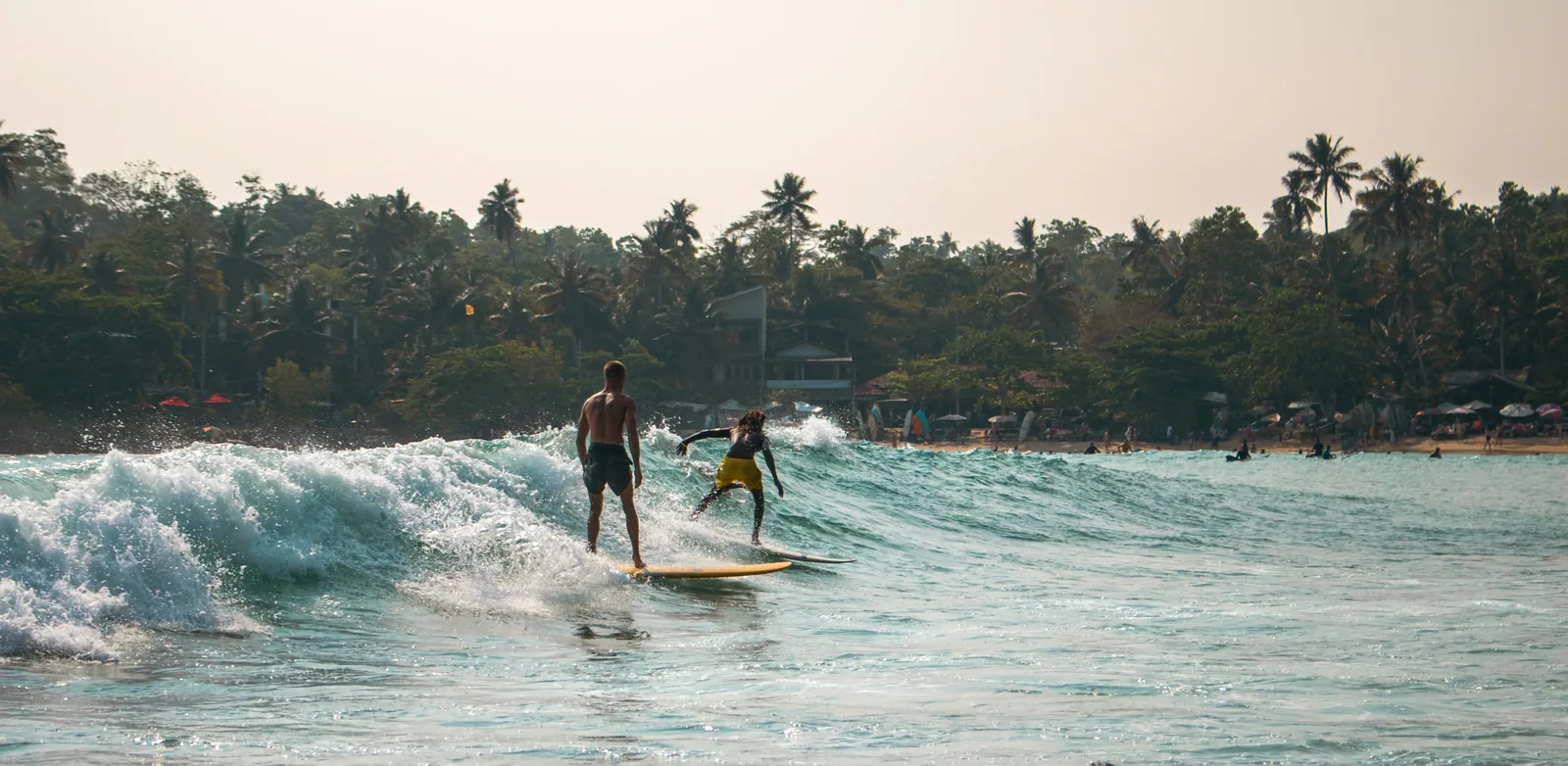 The horseshoe-shaped bay of Hiriketiya beach at golden hour, with surfers in the water and rocky headlands framing the warm orange sky