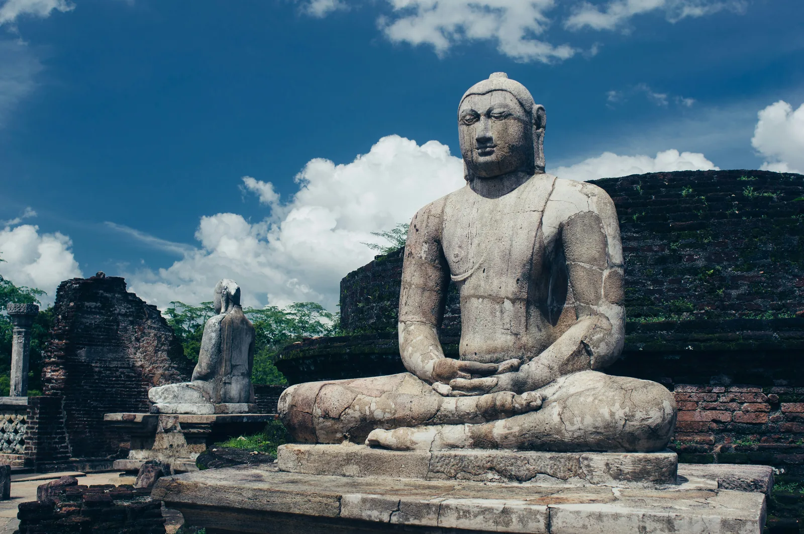 A large seated stone Buddha statue at Gal Vihara, Polonnaruwa, with ancient brick ruins and blue sky behind