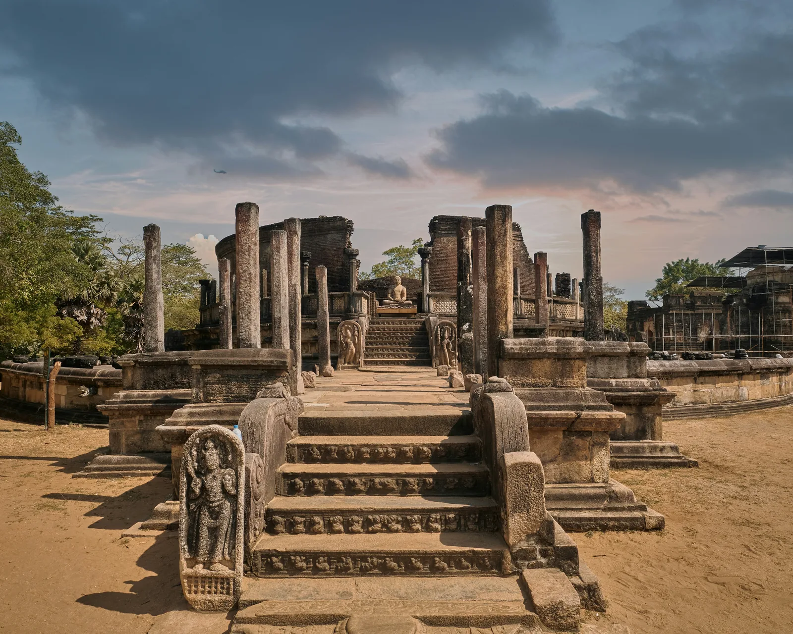 Stone steps and carved pillars of the Vatadage at the Polonnaruwa Quadrangle, with a seated Buddha at the centre and a dramatic cloudy sky