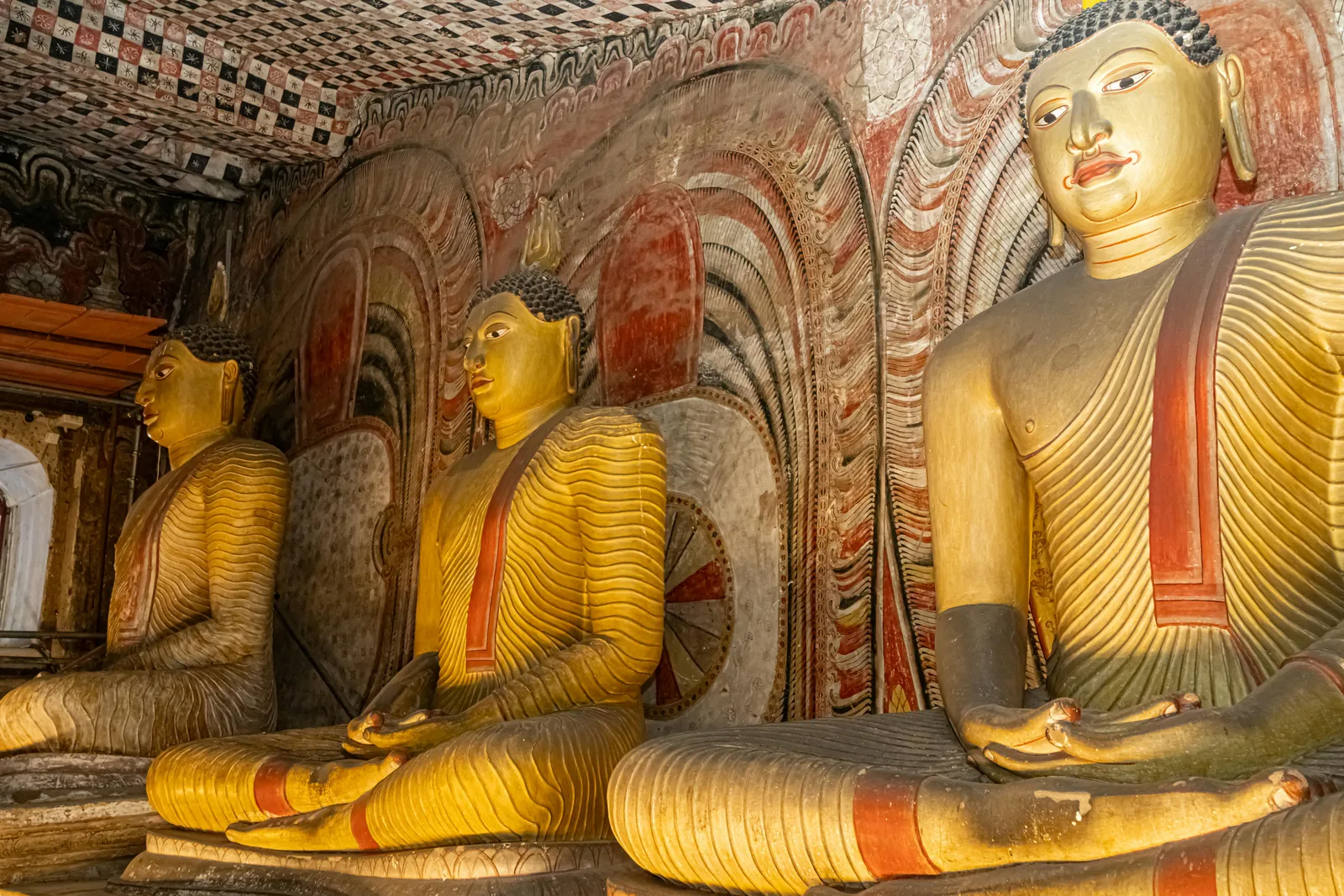 Gilded seated Buddha statues inside the Dambulla Cave Temple, Sri Lanka, with ornate painted frescoes covering the ceiling and walls behind them